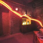 Ein dunkler Raum in dem ein Schrank und eine Schreibmaschine stehen, ein heller Lichstreifen geht durch das Bild.