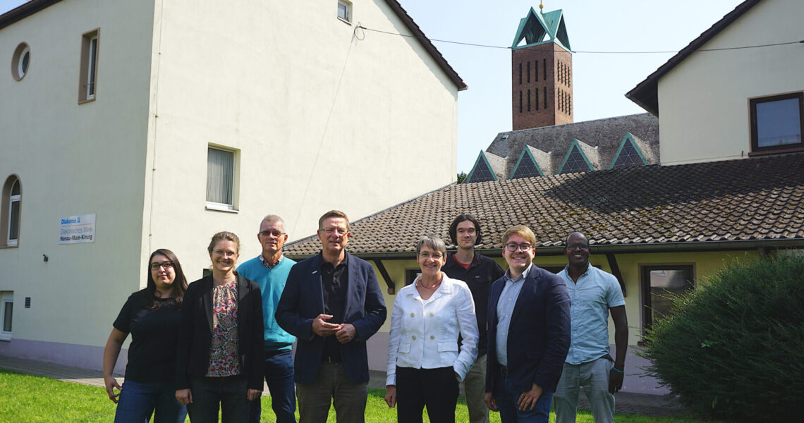 Gruppenfoto von Carsten Tag, Ute Engel, Samuel Mergenthal und den Mitarbeitenden der Flüchtlings- und Migrationsberatung des Diakonischen Werkes Hanau - Main-Kinzig vor dem Gebäude an der Christuskirche.