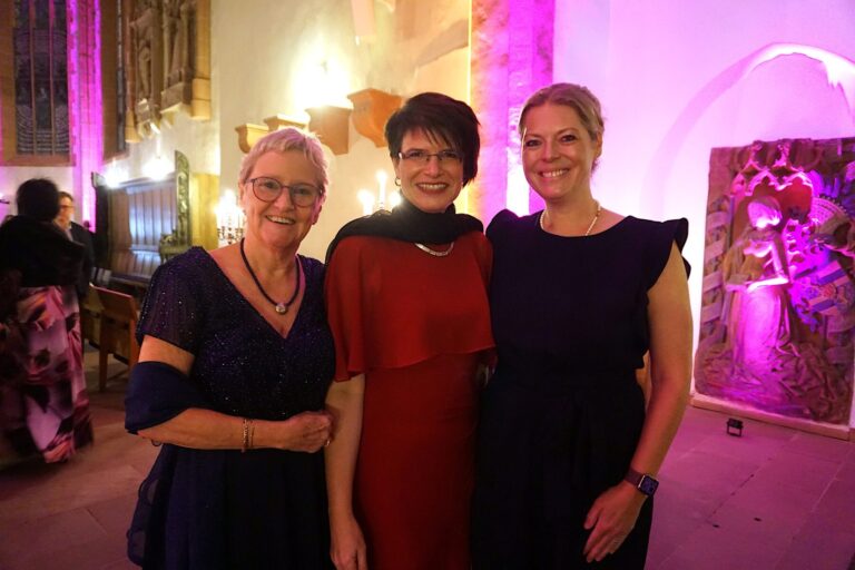 Die drei Organisatorinnen des Balls vor dem stimmungsvoll beleuchteten Chorraum der Marienkirche Hanau.