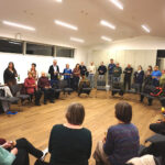 Personen stehen und sitzen in einem Kreis, die Vorsitzende des KV spricht zu den Anwesenden