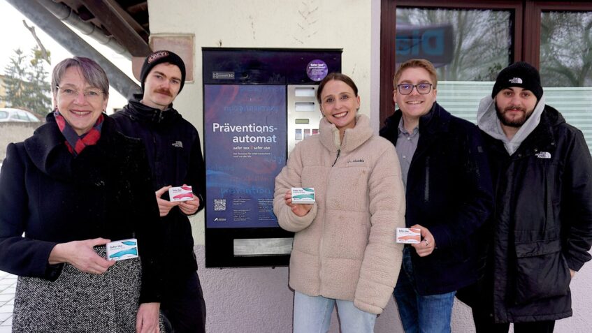 Vertreter von Diakonie und AIDS-Hilfe stehen vor dem neuen Präventionsautomaten, sie halten Päckchen mit Safer Use-Materialien in der Hand.